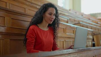 Smiling happy positive Latina woman student girl female typing laptop online chatting using net internet class lesson preparing to exam in university college academy auditorium studying with computer video
