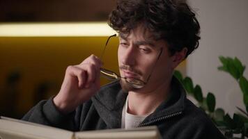 Pensive focused Caucasian guy hipster holding book notebook man reading interesting literature novel hobby leisure reader learn knowledge concentrated male putting on eyeglasses think at home library video