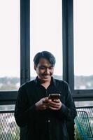 Smiling Asian man using smartphone near a window for communication photo