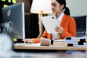 A woman in an orange jacket is sitting at a desk with a tablet photo