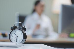 A clock on a desk in front of a woman in the background photo