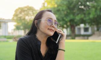 Side view of Asian woman sit on bench in spring park outdoors and look aside while talking by mobile. photo