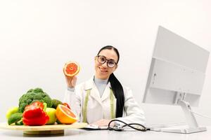 A nutritionist in a lab coat with an avocado in hand sits at a table of fresh fruits and vegetables in a bright office of a modern clinic photo