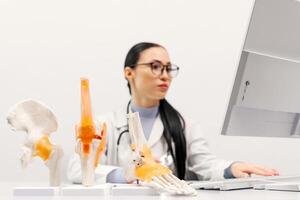 An orthopedic specialist works in a modern office using a computer. Models of the knee joint, pelvic floor and foot are on the table. Concept of orthopedic diagnostics. White background photo