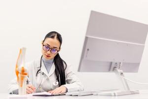 A female physical therapist sits at a table at a computer next to anatomical training models of joints for demonstration. Health Concept photo