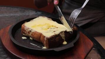A cheesy, buttery toast is cut with a fork and knife on a black skillet, oozing cheddar over golden brown bread. A delectable, appetizing savory meal. video