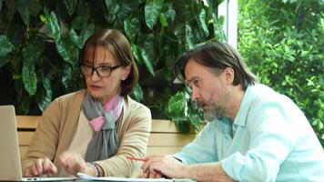 Business Talk. Middle aged man and senior woman collaborate in cafe. Discuss ideas while working on a laptop suggesting New Business or Teamwork or Mentorship and modern work from anywhere lifestyle video