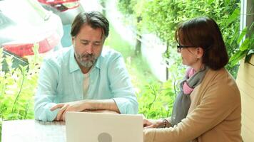 Business Talk. Middle aged man and senior woman collaborate in cafe. Discuss ideas while working on a laptop suggesting New Business or Teamwork or Mentorship and modern work from anywhere lifestyle.. video