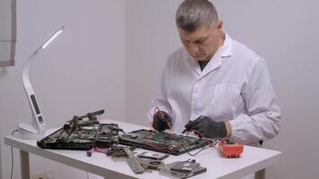 Senior technician in a white lab coat and black gloves carefully using a multimeter to check the circuitry of a disassembled laptop on a tidy white table, surrounded by various electronic parts video