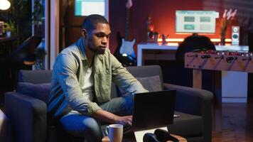 African american guy does research by reading articles and data driven papers in the living room, using online sources to collect information for a new project. Multitasking. Camera B. video