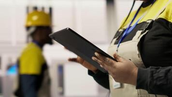Engineer in smart factory oversees robotic assembly lines using tablet app, ensuring quality control during manufacturing. Worker monitoring automated conveyor systems using device, camera B close up video