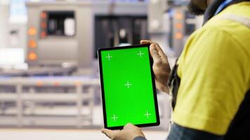 Engineer in smart factory oversees robotic assembly lines using green screen tablet, ensuring quality control. Worker monitoring automated conveyor systems using chroma key device, camera B closeup video