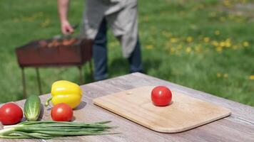 Fresh vegetables on wooden table outdoors. Food for picnic on the blur background of nature. Man frying sausages on a brazier. video