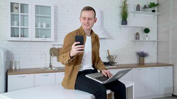 Portrait of a student with a laptop and a phone. Smiling young man is sitting on a kitchen table and using a laptop and mobile phone at home. Quarantine concept. video