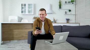 Smiling freelancer with a phone on kitchen background. Young man student using a laptop and mobile phone studying online. Guy working from home enjoying distant job. video