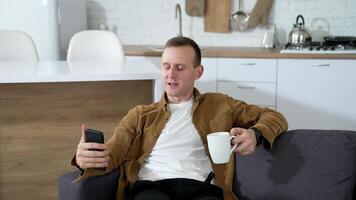 Handsome man using a phone at home. Guy rests on sofa with a cup of coffee and doing photos on a smart phone in the kitchen. video