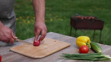 Man cutting radish for a barbeque. Fresh vegetables on the table on the background of a brazel with grilling meat. Time for a picnic. video
