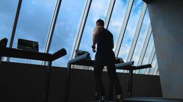 Sportsman on a treadmill indoors on sky background. Fitness guy is running on a simulator in the gym and looking through windows. Rear view. video