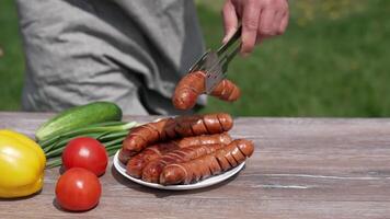 Picnic food on blur nature background. Plate with roasted sausages and fresh vegetables on table. Man adds grilled sausage using tongs to a plate. Close-up. video