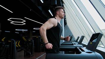 Sportsman is running on a treadmill. Young bodybuilder exercising on a simulator on the background of modern interior of fitness center. Profile view. video