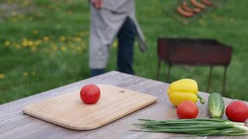 Fresh vegetable on the table in the open air. Tomatoes, cucumbers, onion and pepper on table on the background of a man cooking sausages on grill. video