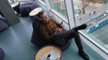 Young businessman sitting at break time. Smiling man sitting by the window and looking into the phone on the city background through the window. Top view. video