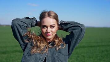 Attractive girl posing on camera outdoors. Portrait of a young woman with long hair in denim jacket looking on camera on green field background. video