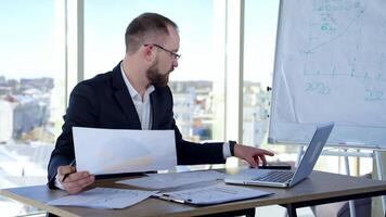 Serious businessman in suit working in office. Entrepreneur sitting at table using laptop, phone and project papers on window background. video