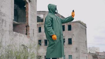 Human in a protective suit and gas mask with a phone. Unrecognizable person in safety uniform, gloves and respirator doing selfie in the destroyed place during radiation. video