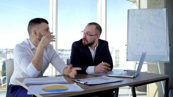 Conversation between two businessman. Successful entrepreneur men sitting at the table in office center and talk happily on window view background. video