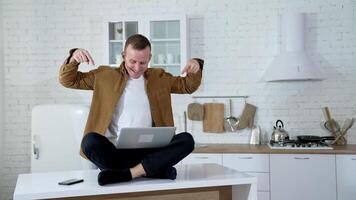 Funny man with a laptop on a table.Smiling freelancer sitting on a table and waiting to press button on a laptop on the kitchen background. video