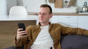 Young guy writing messages on a phone at home. Portrait of a handsome man sitting on a couch with a smartphone in the kitchen. video