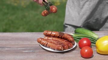 Ready grilling sausages with vegetables on the table outdoors. Chef puts roast sausage into a plate. Tasty meal for a picnic. video