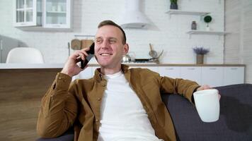 Young man talking the phone at home. Happy guy resting with a cup of coffee and having a pleasant phone conversationin the kitchen. video