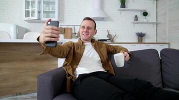 Young man taking photo of himself at home. Handsome guy sitting on sofa using mobile phone and drinking tea indoors. Smiling man enjoying sitting at home during quarantine. video