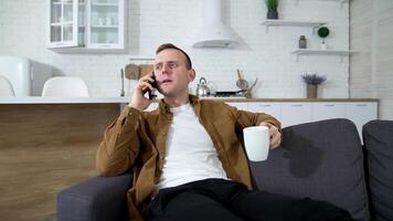 Guy with a mobile phone in the kitchen. Young man talking the phone while sitting with a white mug on a couch at home. video