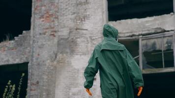 Perplexed survivor in abandoned place. Human in safety uniform and gas mask standing alone and looking to the sides on the background of destroyed buildings. video