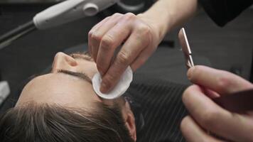 Using straight razor with a sharp blade, barber carefully removes stubble from client's cheek, then applies soothing lotion with a cotton pad. video