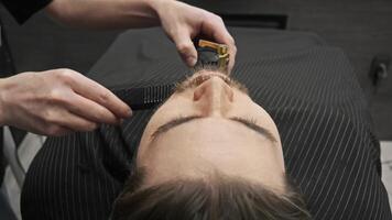 To achieve an even length for the beard, the hairstylist cuts the client's hair using a comb and a shaving machine. Close up. High angle video