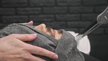 Young man's face is wrapped in warm, damp towel as he lies under steam machine, which creates suitable temperature to soften skin and make it easier to shave in barber's shop. video