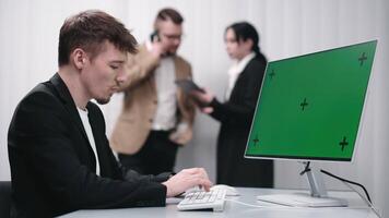 IT technician working at a desktop computer with a Chroma Key green screen, while colleagues brainstorm in the office. Concept e-commerce development, marketing video