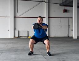 A plus-size man performing kettlebell squats in an empty garage with a rugged, industrial light vibe. photo