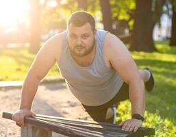 A plus-size man using a park bench to do push-ups under the afternoon sun, wearing comfortable sportswear. photo