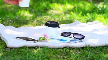 A cozy spring scene with glasses, a notebook, and a cherry blossom twig on a blanket in the grass. A hand reaches for a coffee cup, video