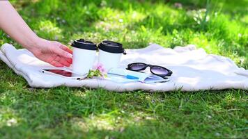 A cozy spring scene with glasses, a notebook, and a cherry blossom twig on a blanket in the grass. A hand reaches for a coffee cup, capturing a peaceful outdoor moment. video