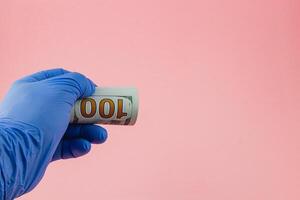 Hand in blue glove holds rolled 100 bill on pink bg photo