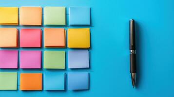 Colorful sticky notes arranged in neat rows on blue surface with black pen casting soft shadow, creating organized workspace feel photo