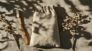 Top view of a bamboo toothbrush and a small linen bag with leaf shadows and dried flowers on beige fabric. Eco-conscious lifestyle photo