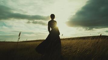 Lonely woman in a long dress standing in the windy field with sun rays breaking through the clouds photo