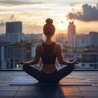 Young woman meditating on balcony at sunset, sitting in lotus pose with city skyline view, relaxing evening session photo